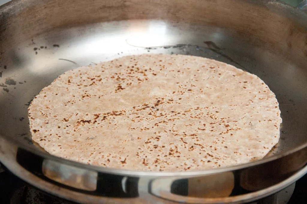 A rolled whole wheat chapati cooking in a pan, with small brown spots across the surface