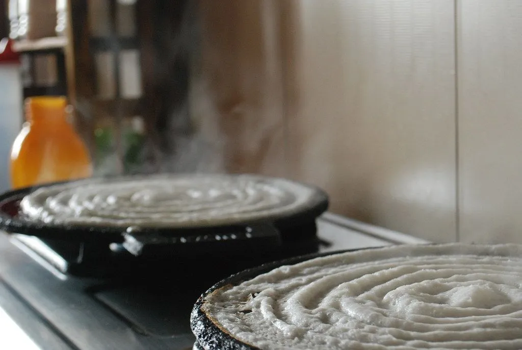 Dosa batter spread in rings on two dark tawas over a flame, with steam rising