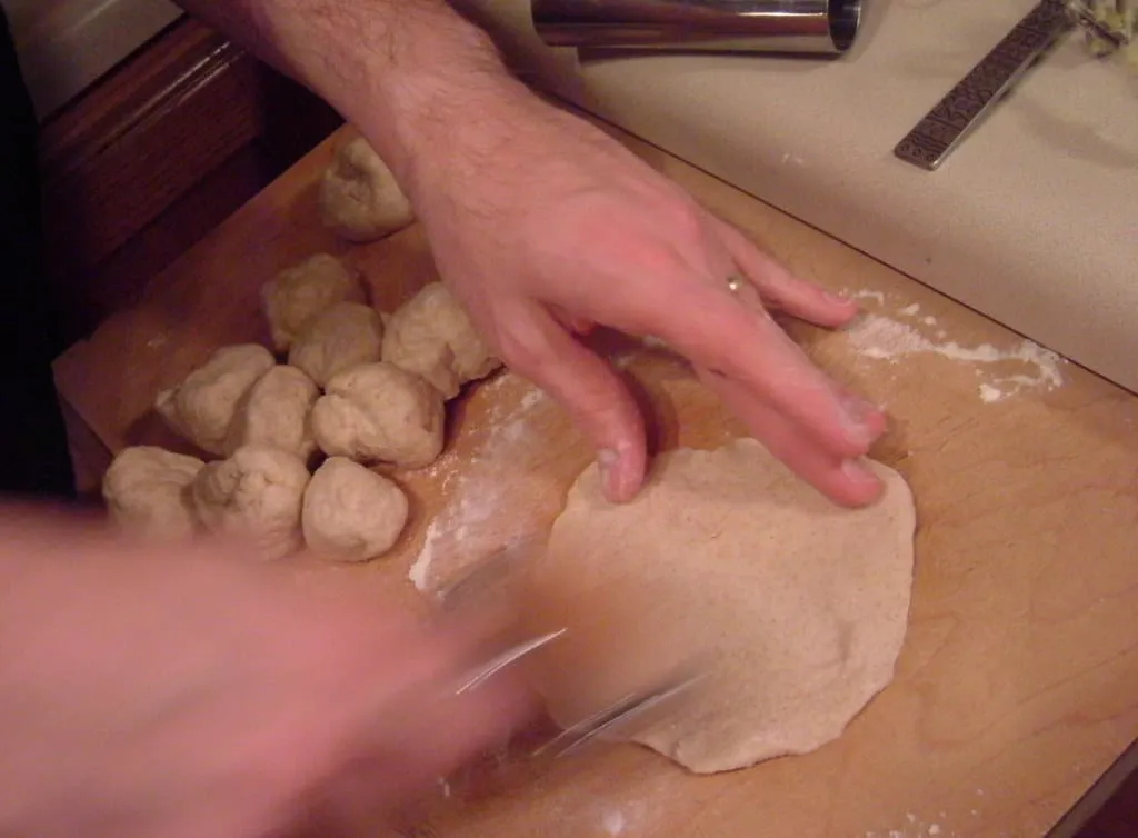 A hand rolling out a small round of whole wheat dough on a wooden board, with balls of dough waiting beside it