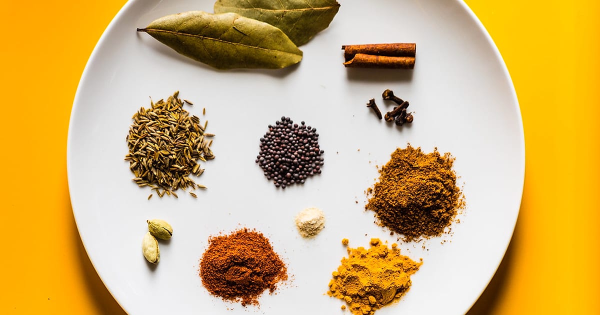 Whole and ground Indian spices — bay leaf, cinnamon, cloves, cumin, mustard seeds, cardamom, chilli, turmeric and garam masala — on a white plate