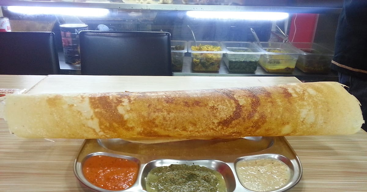 A long, crisp golden ghee dosa resting across a steel plate of tomato, mint and coconut chutneys