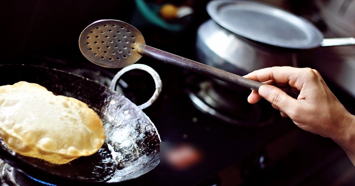A puri puffing up in hot oil in a dark iron kadai, a slotted spoon ready to lift it out