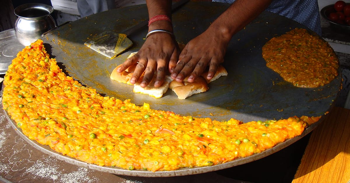 A street cook toasting pav on a huge iron tawa ringed with bright orange pav bhaji