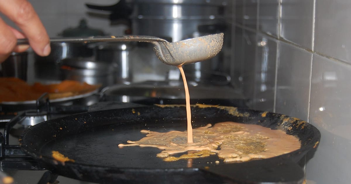 Dosa batter streaming from a ladle onto a hot black tawa, the moment that decides whether the dosa will stick