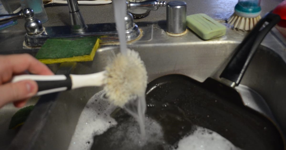 Scrubbing a black cast iron griddle with a brush under running water in the sink