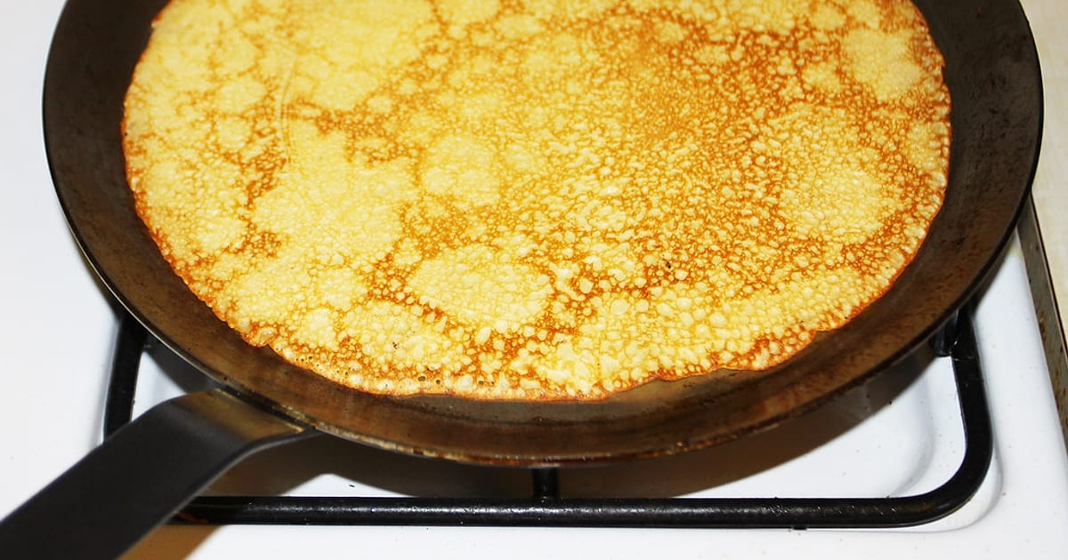 A thin, lacy golden crêpe cooking in a dark, well-seasoned flat iron pan — it releases without sticking