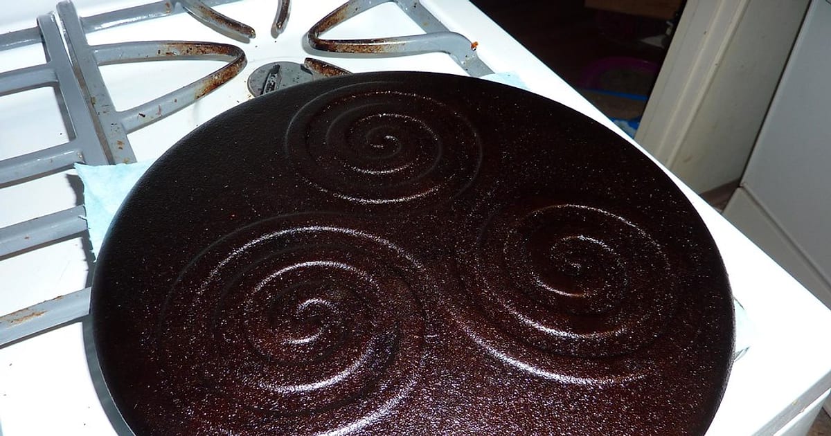 A dark, glossy iron pan with a spiral pattern after its third coat of seasoning, cooling on the stove